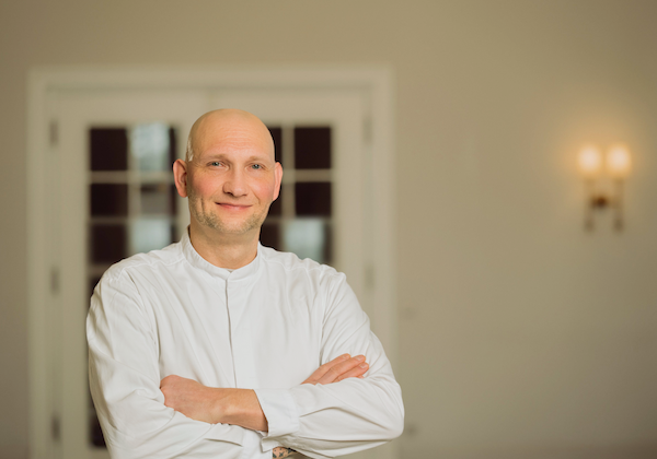 Ein Mann in weißem Hemd mit verschränkten Armen lächelt vor einer Tür.