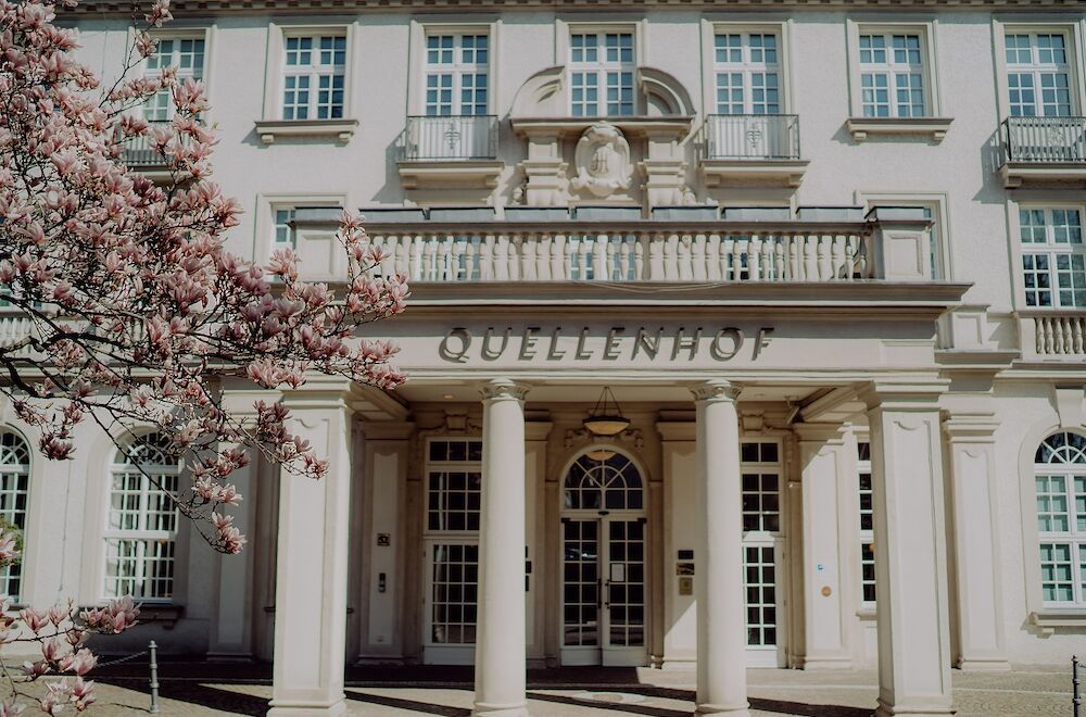 Außenansicht des Hotels „Quellenhof“. Cremefarbenes Gebäude mit Säulen, Balkon und blühendem rosa Baum.
