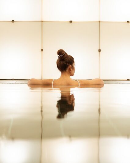 Eine Frau im Pool, die Arme am Beckenrand abgestützt. Die Szenerie ist minimalistisch und dezent beleuchtet.