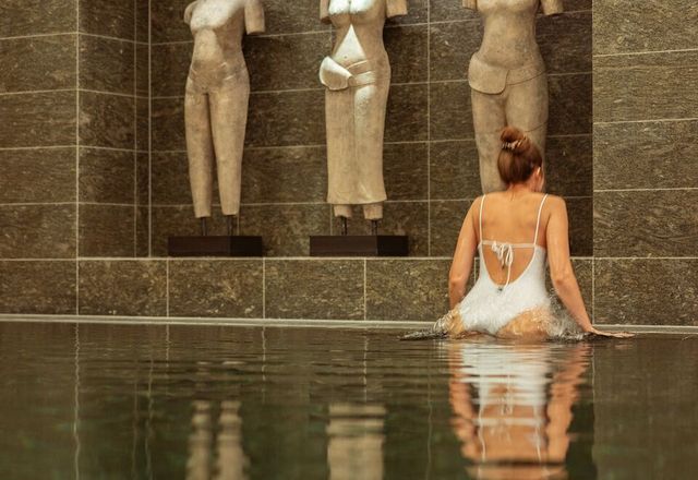 Eine Frau in einem weißen Badeanzug sitzt in einem Pool, im Hintergrund Statuen, dunkle Fliesenwände.