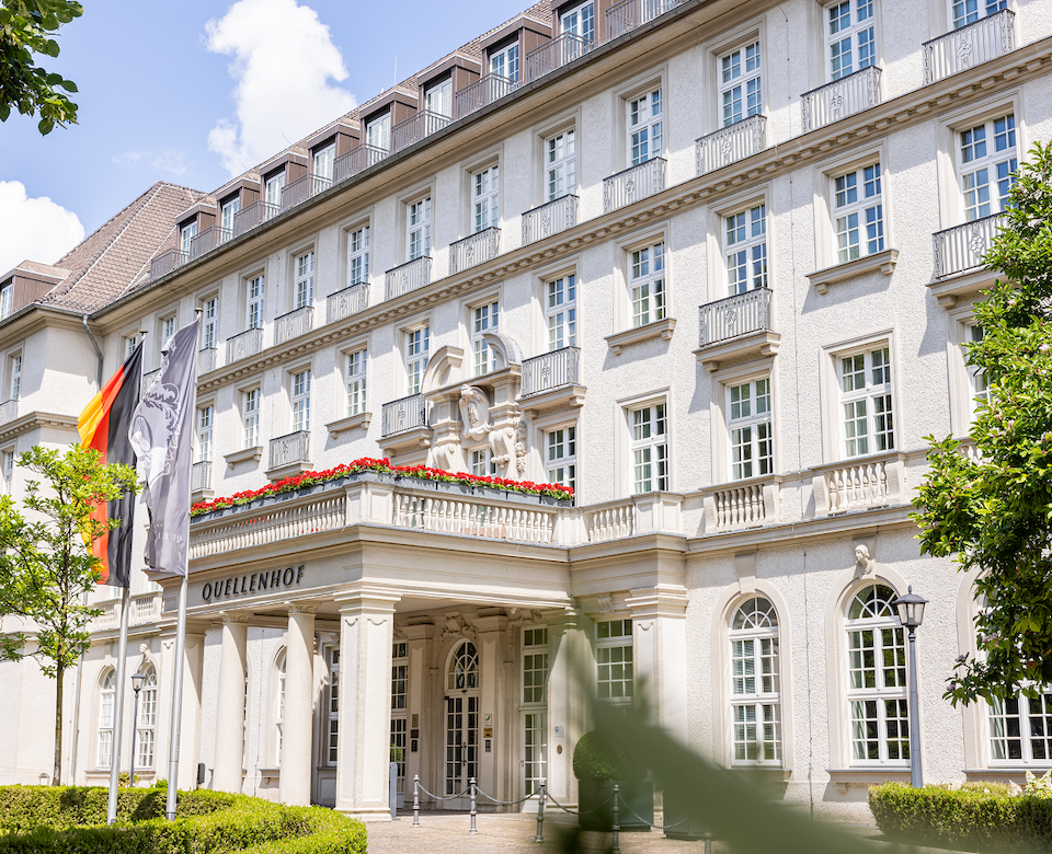 Weißes Gebäude mit mehreren Stockwerken, Balkonen und Fahnen, ein sonniger Tag.