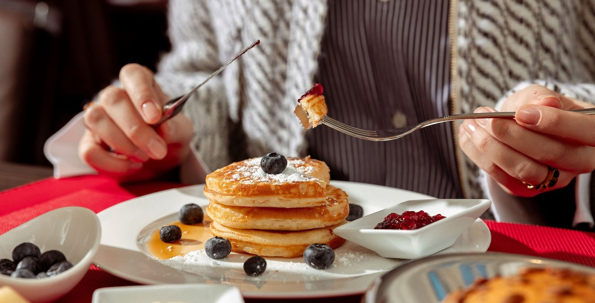 A person is eating pancakes with blueberries, syrup, and jam on a white plate.
