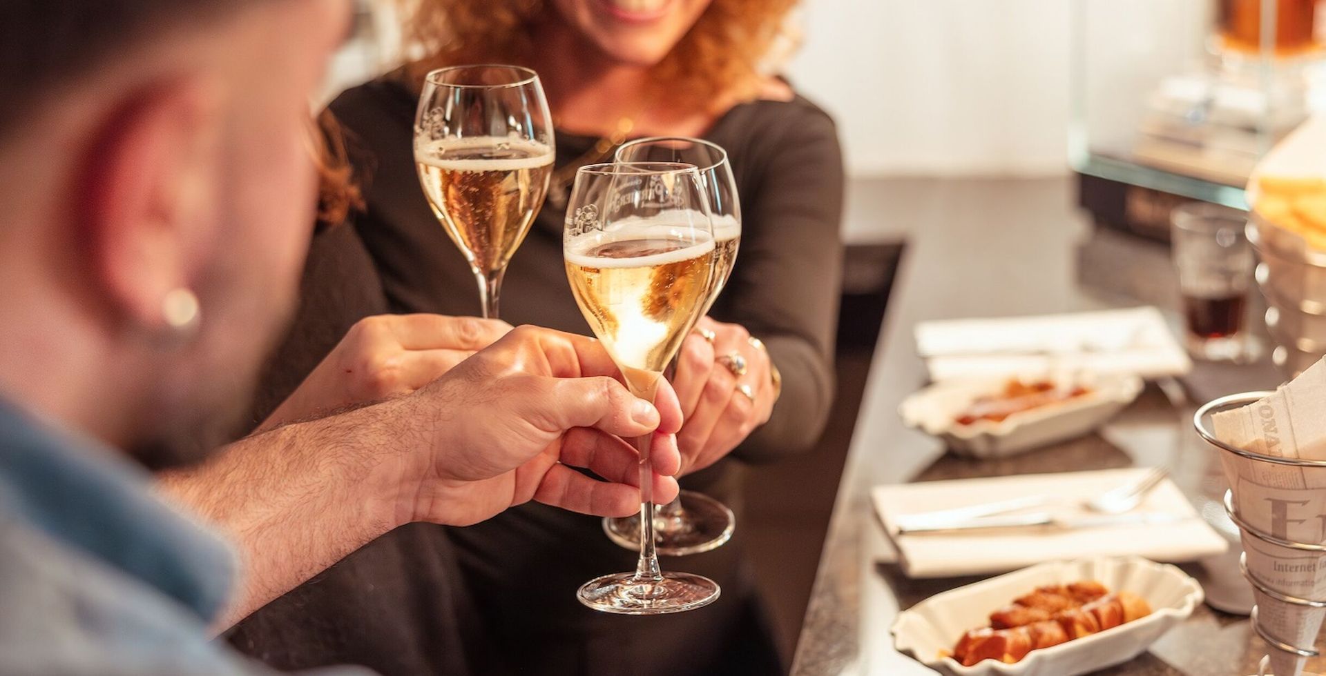 Zwei Menschen stoßen mit Champagnergläsern an; sie halten ihre Getränke in der Hand und lächeln. Im Hintergrund sieht man eine Bar mit Snacks.