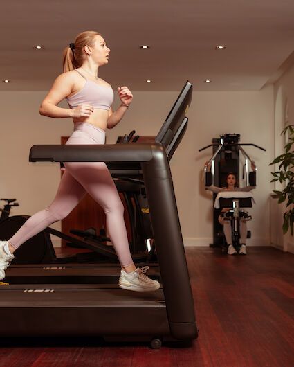A woman is running on a treadmill at a gym; in the background, another person is using a weight training machine.
