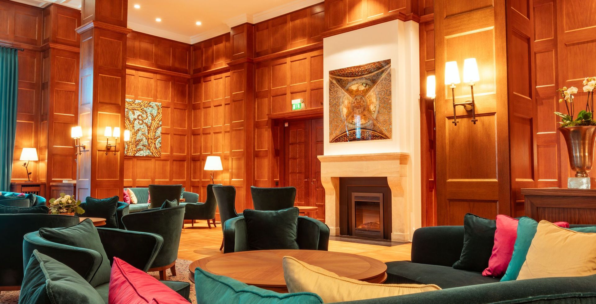 Lounge-Bereich mit Holzvertäfelung, Samtsitzen in Türkis und Dunkelgrün, einem Kamin und sanfter Beleuchtung.