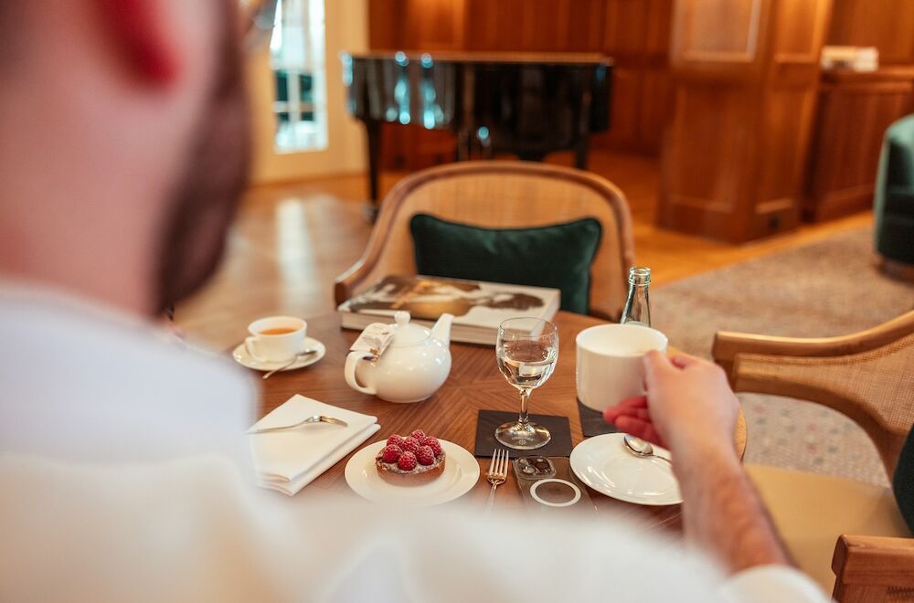 Eine Person sitzt mit Tee und Gebäck an einem Tisch in einem Raum mit einem Klavier.