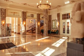 Grandiose Hotellobby mit Marmorsäulen und -böden, Treppenhaus und Kronleuchter. Sonnenlicht strömt herein.