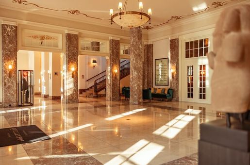 Grandiose Hotellobby mit Marmorsäulen und -böden, Treppenhaus und Kronleuchter. Sonnenlicht strömt herein.