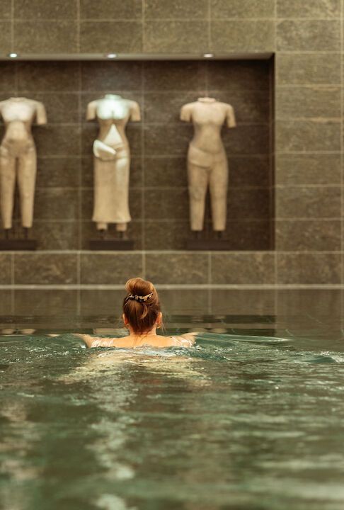 Eine Frau schwimmt in einem Pool und blickt auf Statuen in einer Wandnische. Dunkle Fliesen und neutrale Farbtöne.