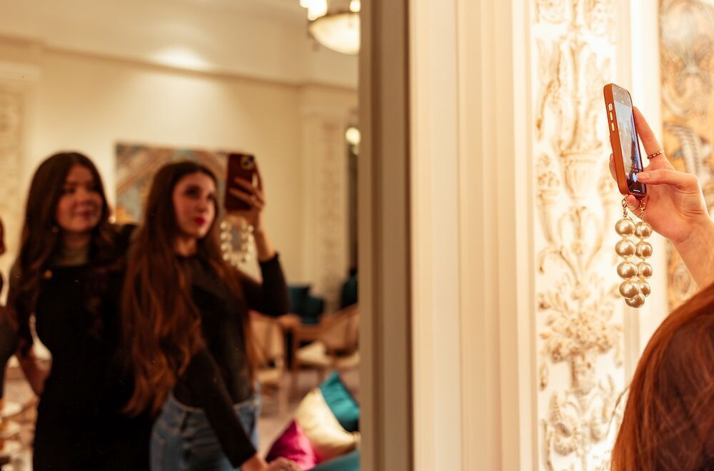 Two women are taking a selfie in the mirror; one is holding the phone. A lavish room, warm lighting.