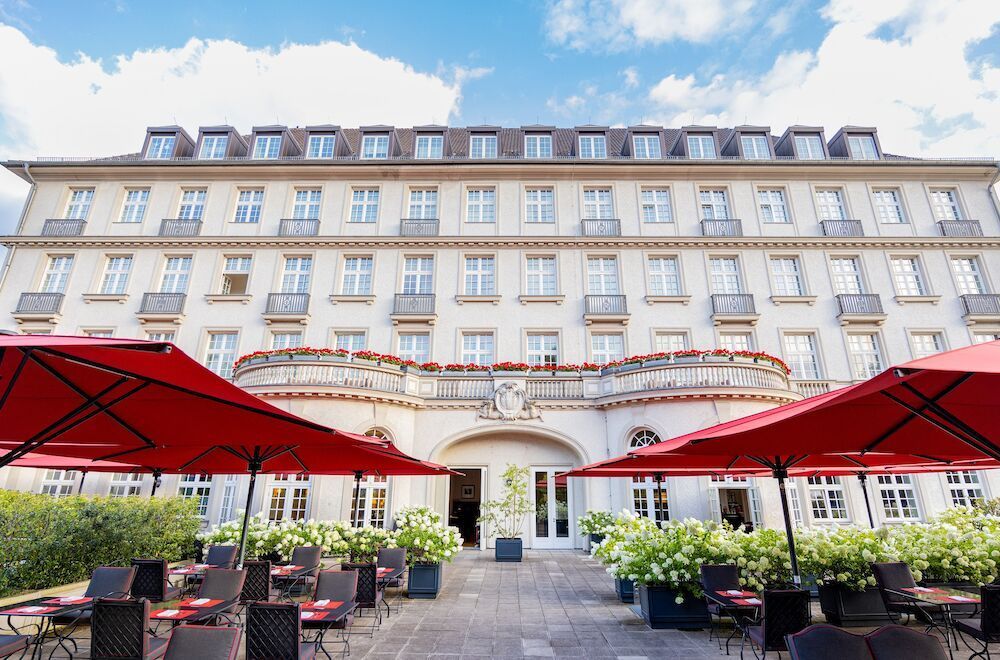 A terrace with red umbrellas and tables in front of a large, bright hotel building.