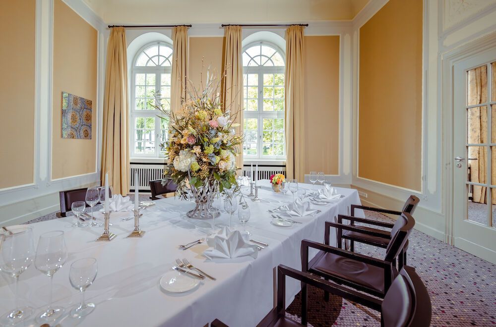Elegantes Esszimmer mit langem, für ein Essen gedecktem Tisch, hohem Blumengesteck in der Mitte und großen Fenstern.