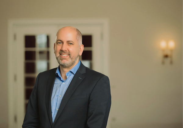 Ein Mann in blauem Hemd und dunklem Anzugjackett lächelt in einem hellen Innenraum.