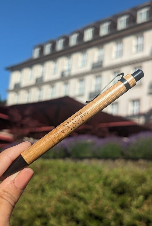 Eine Hand hält einen Bambusstift mit eingraviertem Text vor einem verschwommenen Gebäude und Grünflächen.