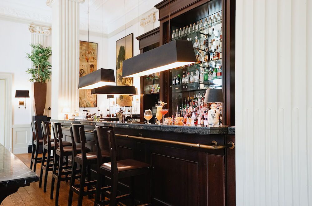 The bar inside is furnished with dark wood, bar stools, and pendant lights; bottles line the shelves.