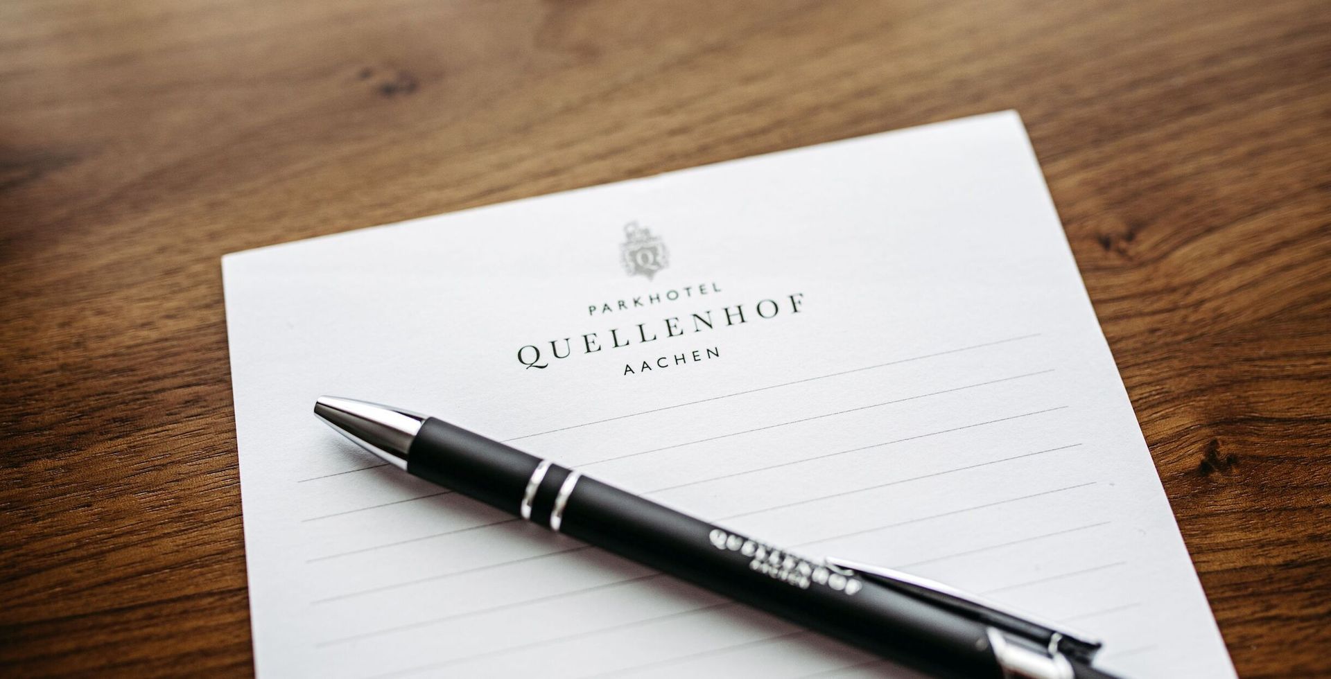 Ein Stift auf einem linierten Notizblock mit der Aufschrift „Quellenhof“ auf einem Holztisch.