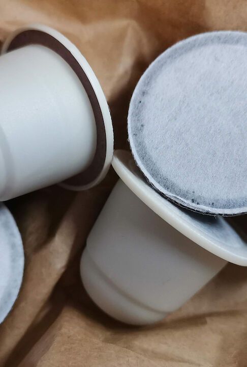 Nahaufnahme von Kaffeekapseln in braunem Papier. Weiße Plastikbecher mit dunklem Filter.