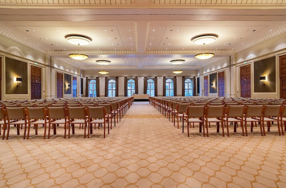 Großer, leerer Ballsaal mit Stuhlreihen vor einer Bühne. Beiger Teppichboden, dekorative Decke und Fenster.