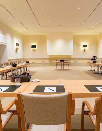 Konferenzraum mit hellen Holztischen und -stühlen, beigen Wänden und einer Projektionsfläche.