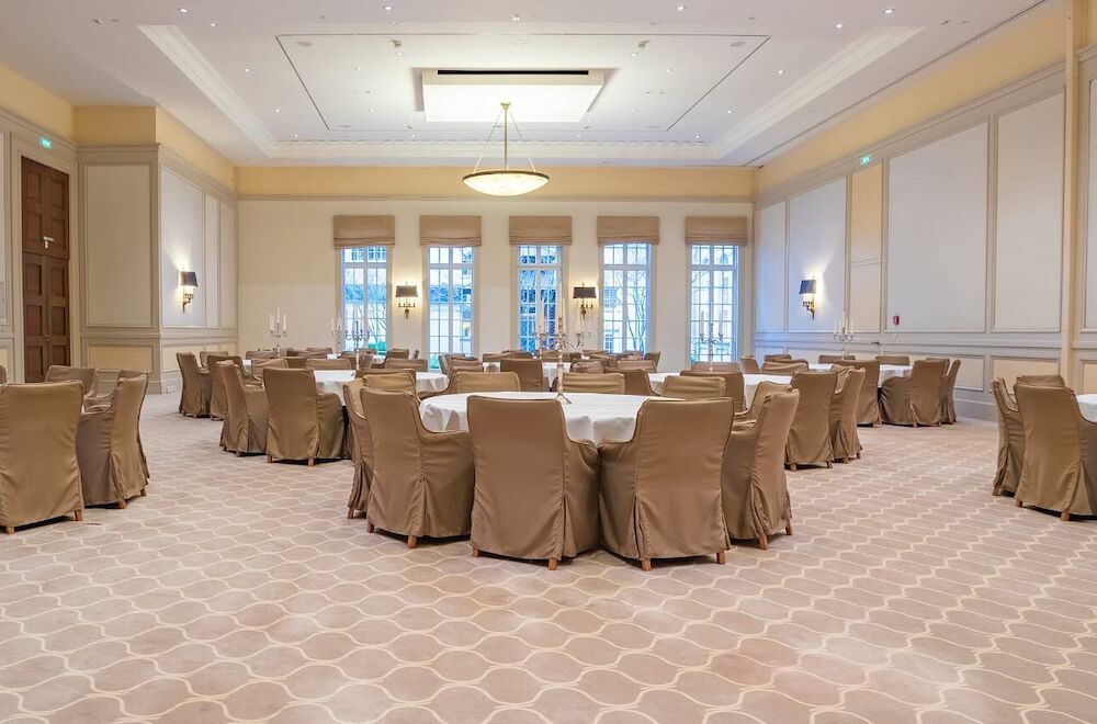 Leerer Festsaal mit runden Tischen, gepolsterten Stühlen, beigefarbenem Teppich und dekorativen Wandpaneelen.