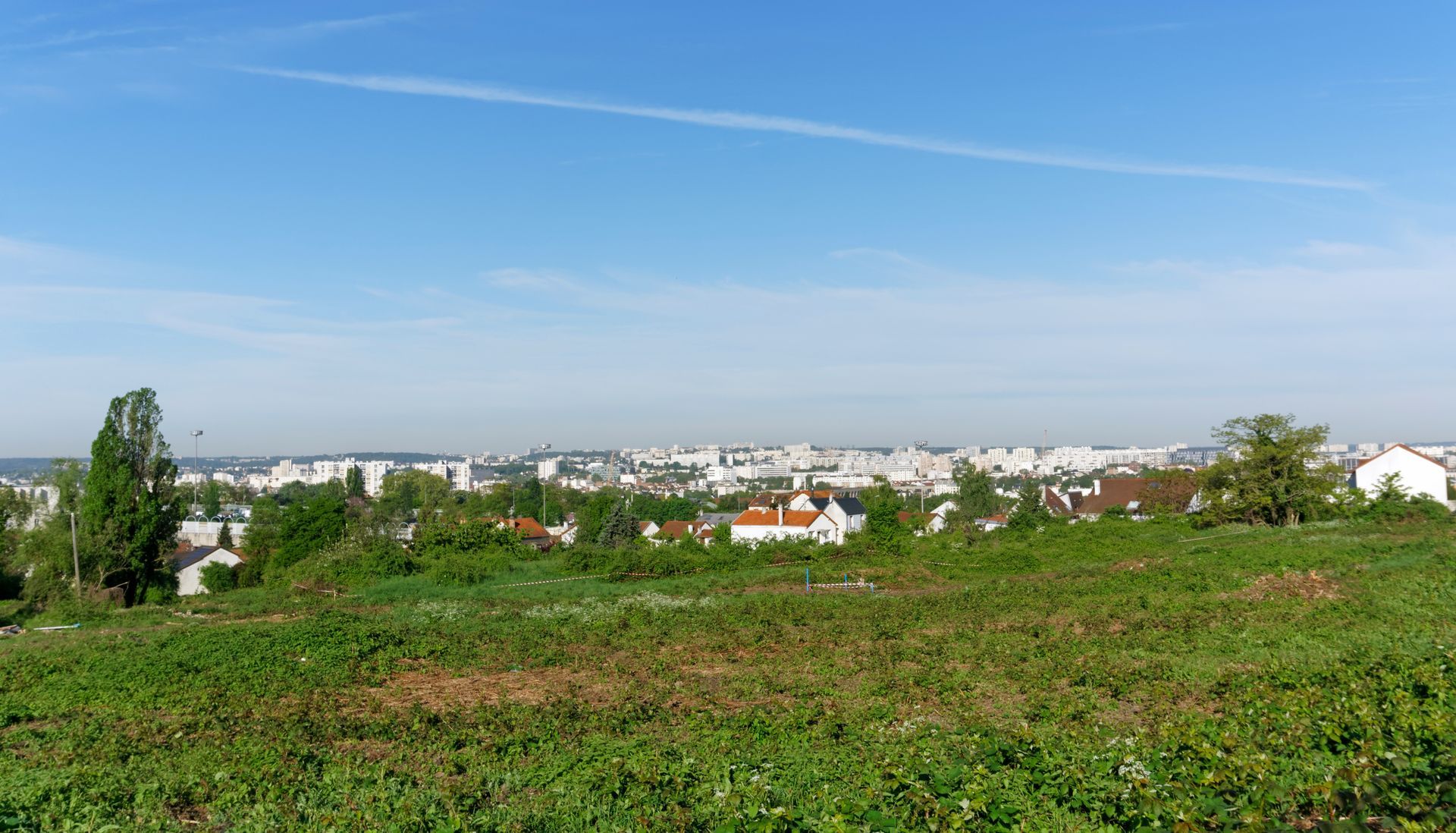 Champ verdoyant au premier plan, maisons et bâtiments au loin sous un ciel bleu.