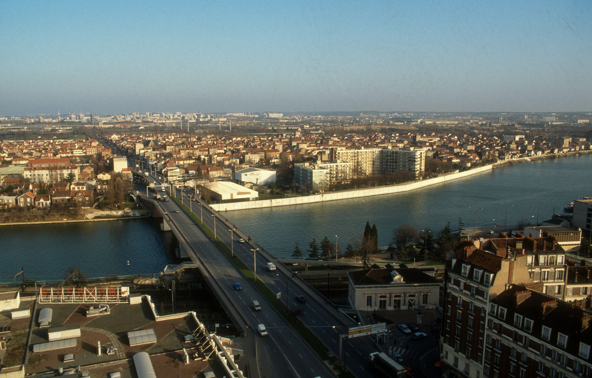 Vue urbaine avec un pont enjambant une large rivière, des bâtiments bordant les rives sous un ciel bleu.