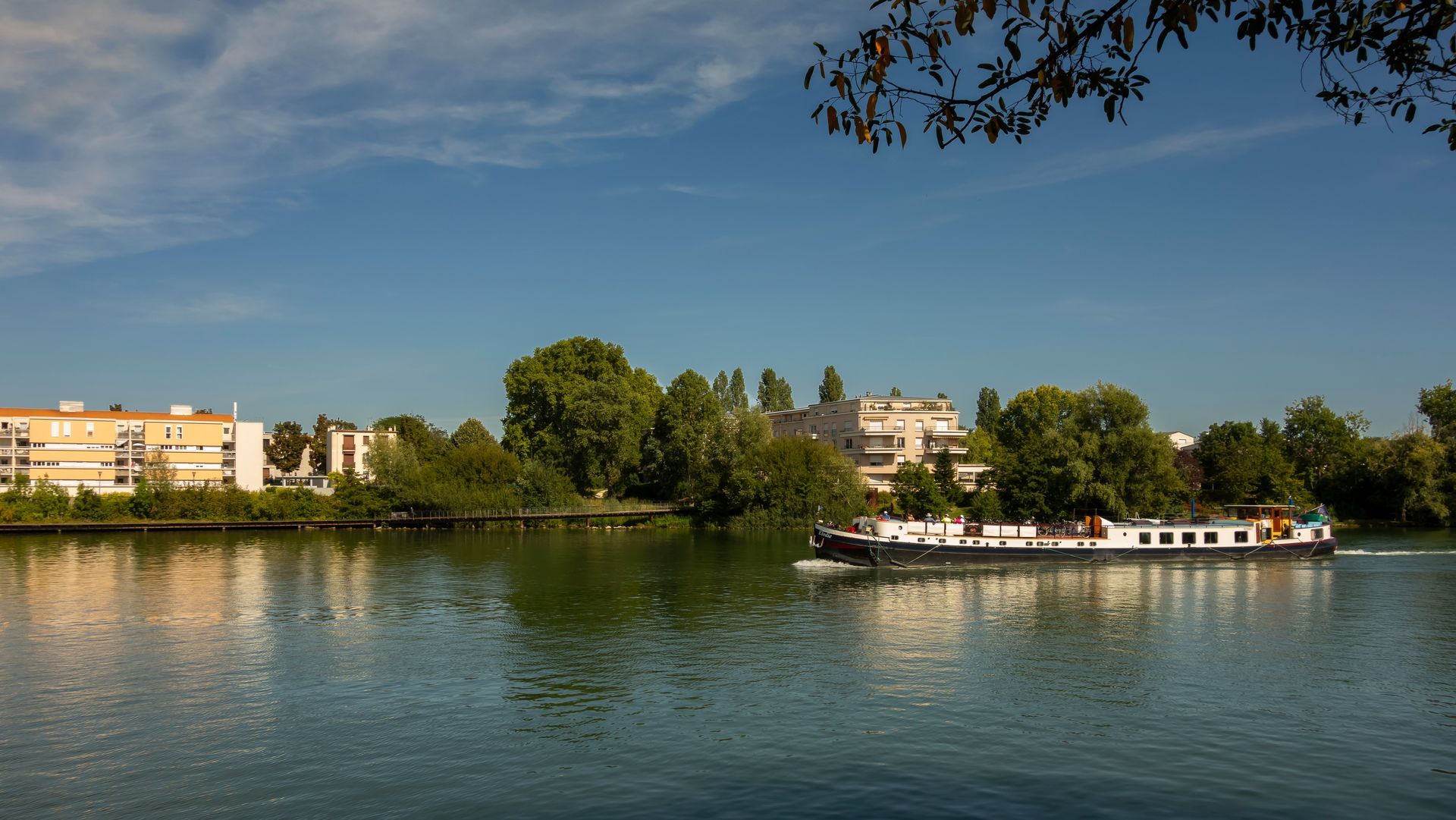Une péniche glisse sur une rivière calme sous un ciel bleu, bordée de bâtiments et d'arbres.