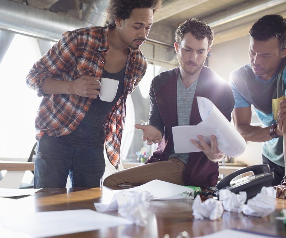 Trois hommes travaillent ensemble à une table, en consultant des documents ; l'un d'eux tient une tasse.