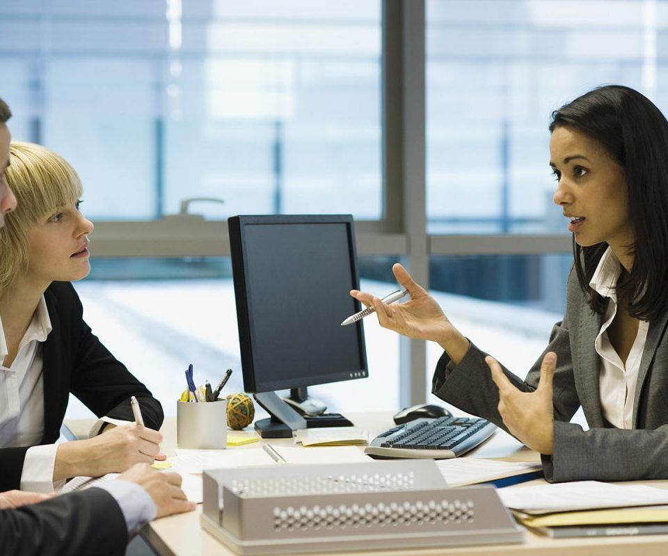 Une femme en tailleur fait des gestes en parlant à deux collègues dans un bureau.