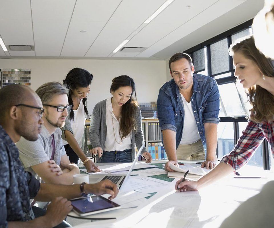 Un groupe de personnes collaborent autour d'une table avec un ordinateur portable, des documents et une tablette, dans une pièce bien éclairée.