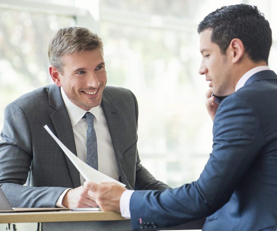 Deux hommes en costume, assis à une table, examinent des documents ; l'un sourit, l'autre est au téléphone.