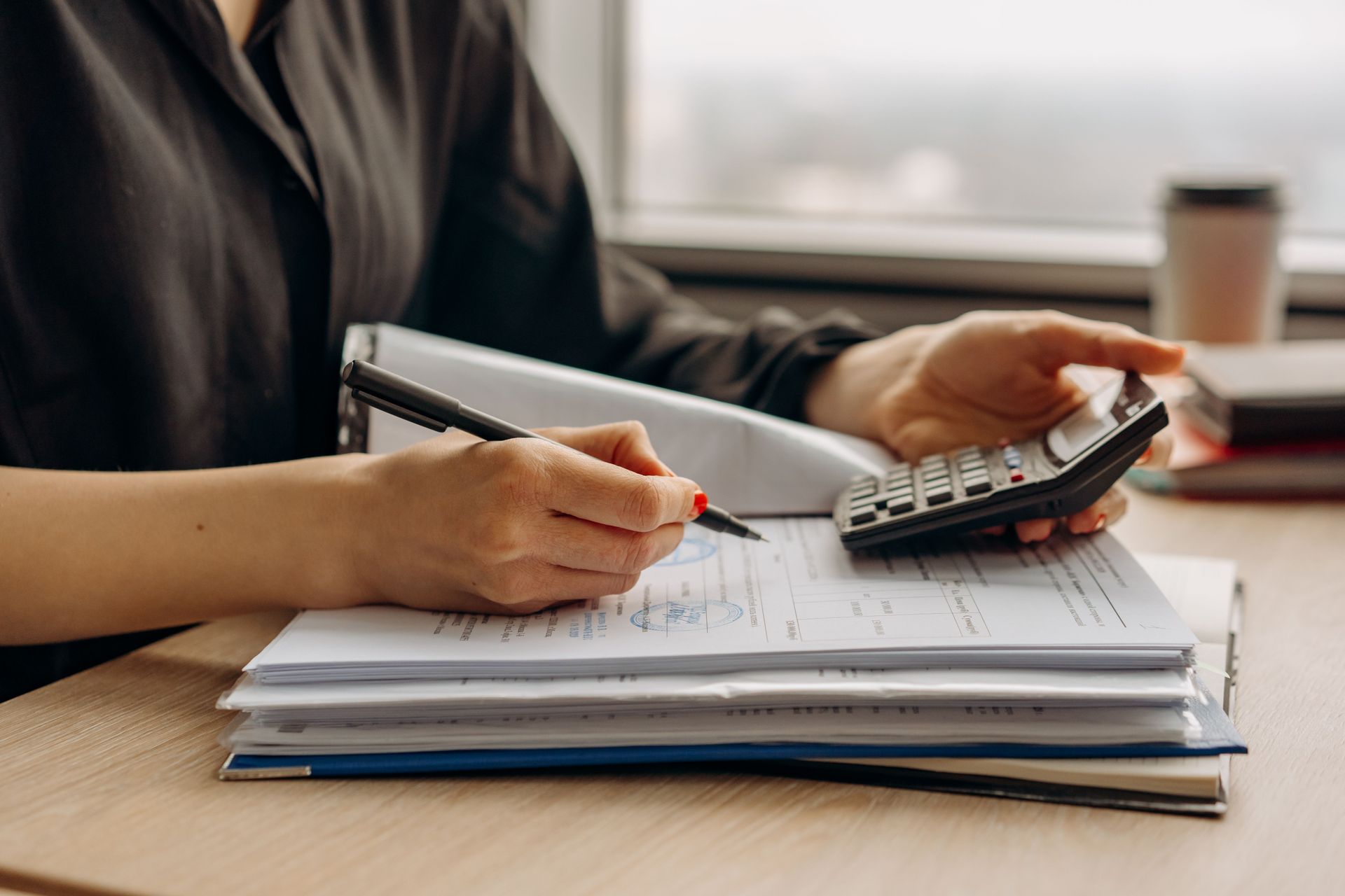 Des mains écrivant sur des papiers tout en tenant une calculatrice au-dessus d'une pile de documents sur un bureau.
