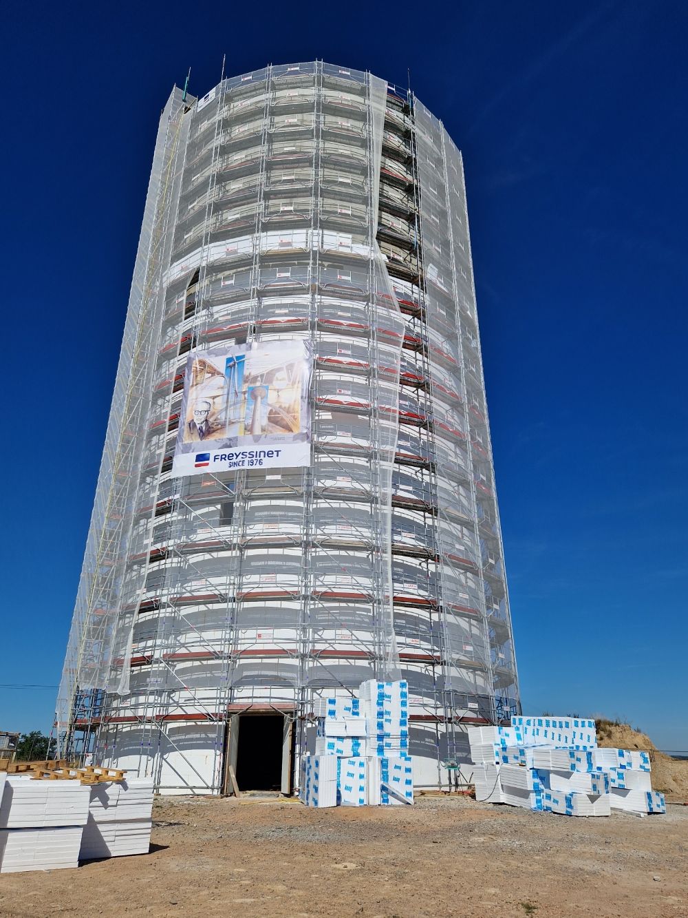 Une tour cylindrique en rénovation, enveloppée d'échafaudages blancs et de treillis de construction, se détache sur un ciel bleu clair.