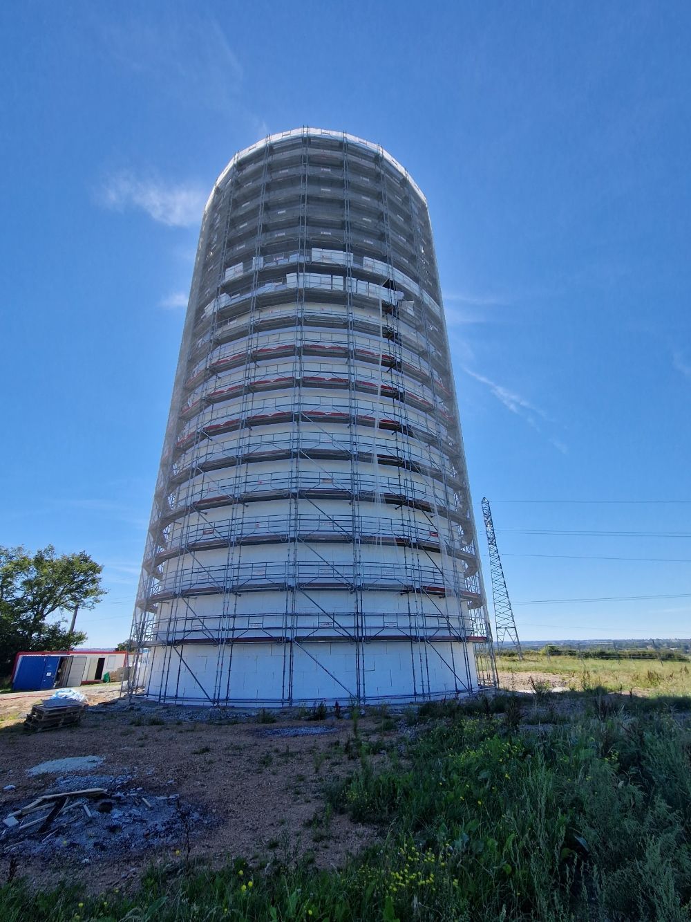 Une haute tour cylindrique en béton, enveloppée d'échafaudages blancs, se dresse sous un ciel bleu limpide, entourée d'un champ ouvert.
