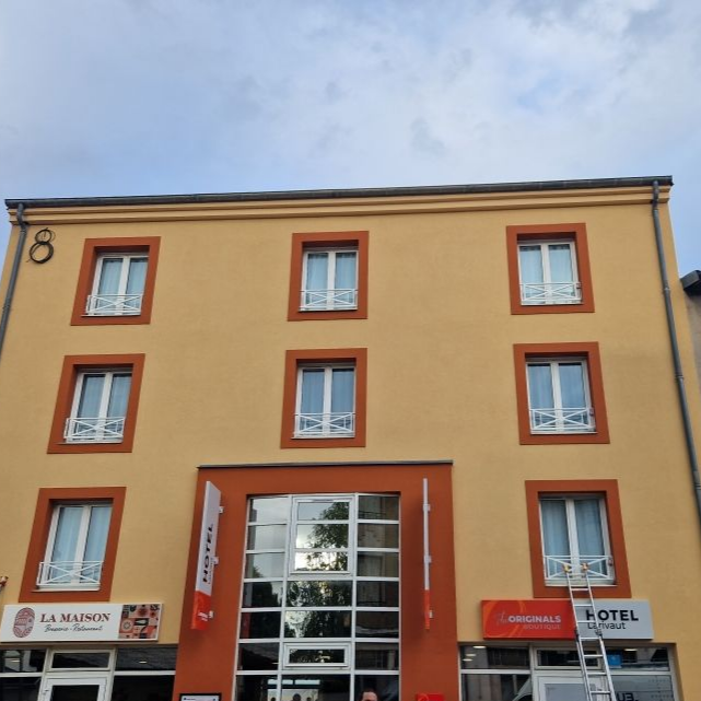 Un hôtel de trois étages couleur beige avec des finitions orange et des encadrements de fenêtres blancs, comprenant des commerces au rez-de-chaussée et une entrée vitrée.