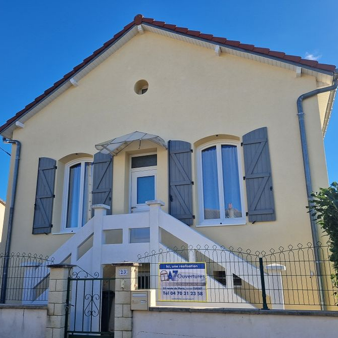 Une maison couleur crème avec des volets gris et un escalier blanc menant à la porte d'entrée, sous un ciel bleu limpide.