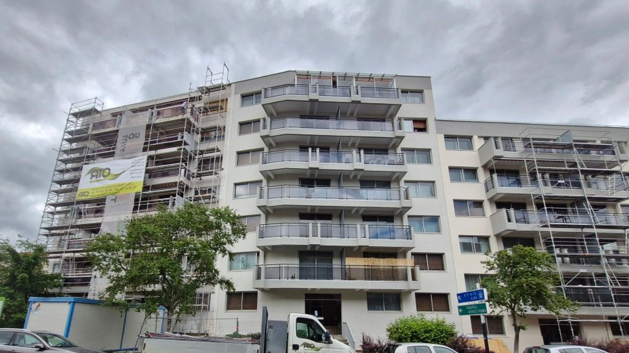 Un immeuble d'appartements en rénovation, avec des échafaudages de chaque côté, une façade blanche et des balcons, sous un ciel nuageux.