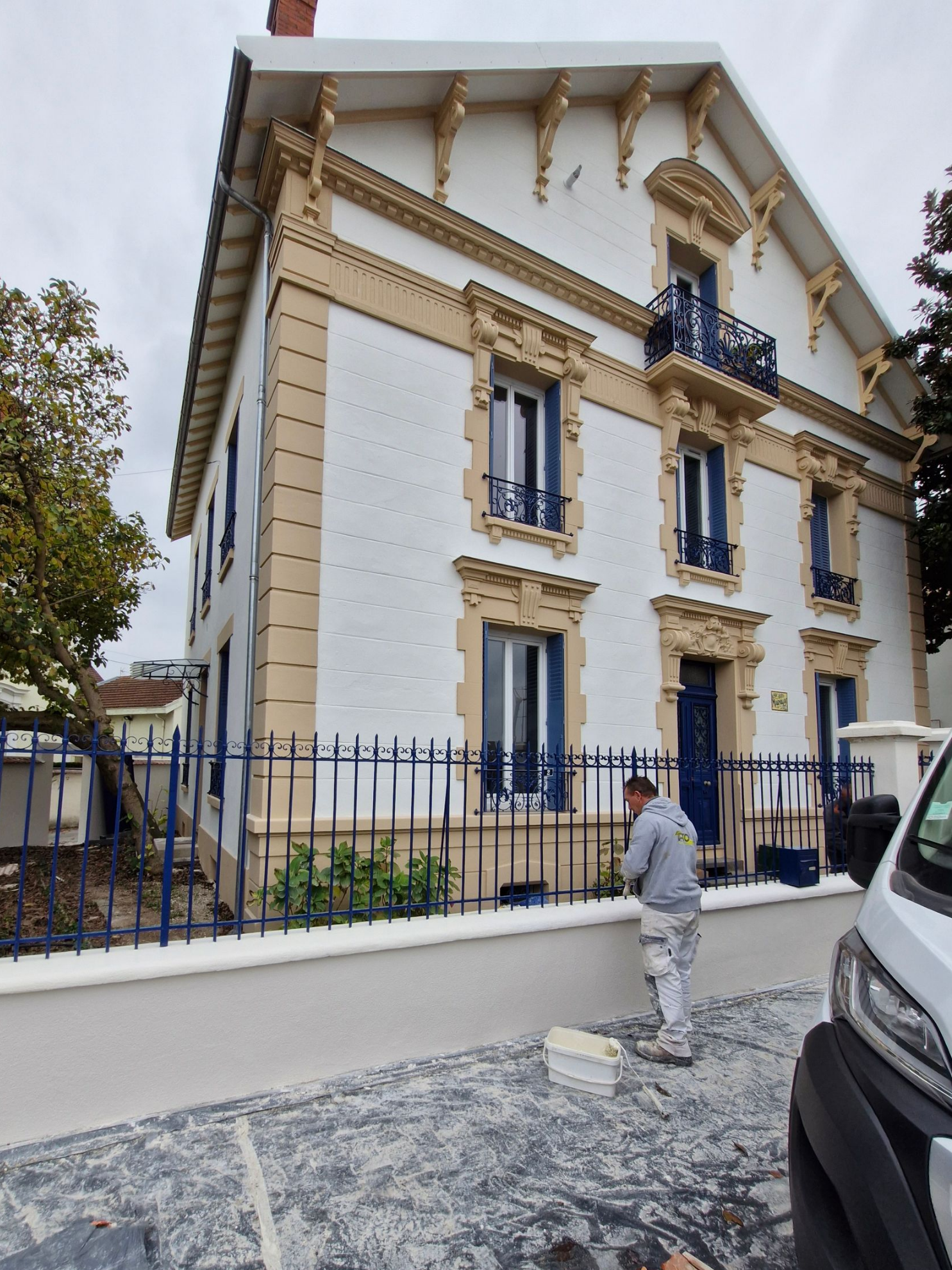 Un ouvrier peint une clôture métallique bleue devant une maison historique blanche de trois étages ornée de boiseries beiges.