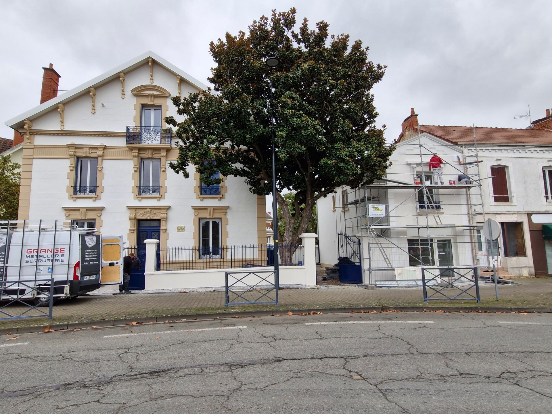 Deux immeubles mitoyens de plusieurs étages, l'un avec des échafaudages et un grand arbre central, bordent une rue asphaltée.