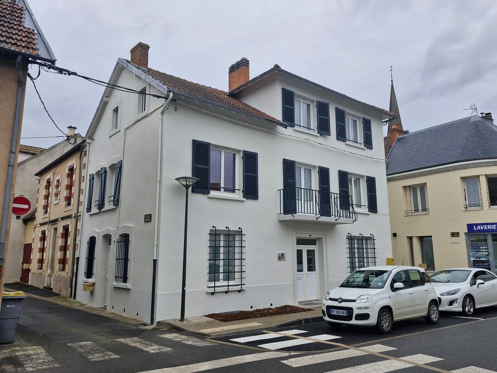 Un immeuble résidentiel blanc de trois étages aux volets sombres, situé à un coin de rue, avec deux voitures garées devant.