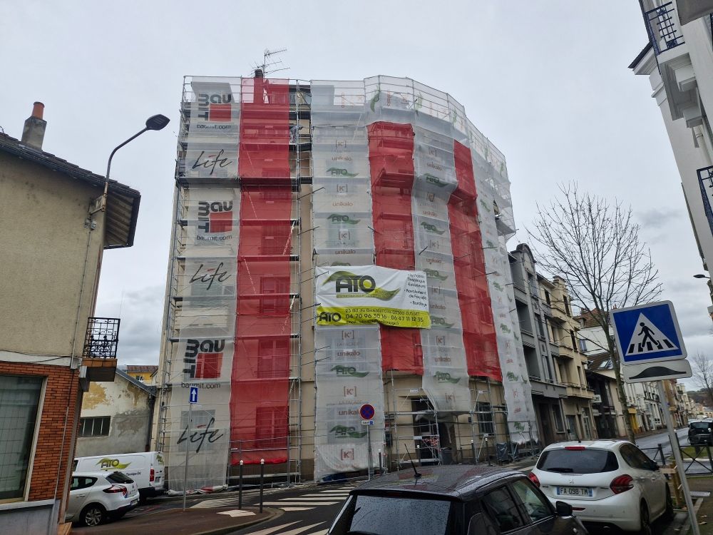 Un immeuble de plusieurs étages en rénovation, recouvert de filets de sécurité blancs et rouges, situé à un coin de rue.