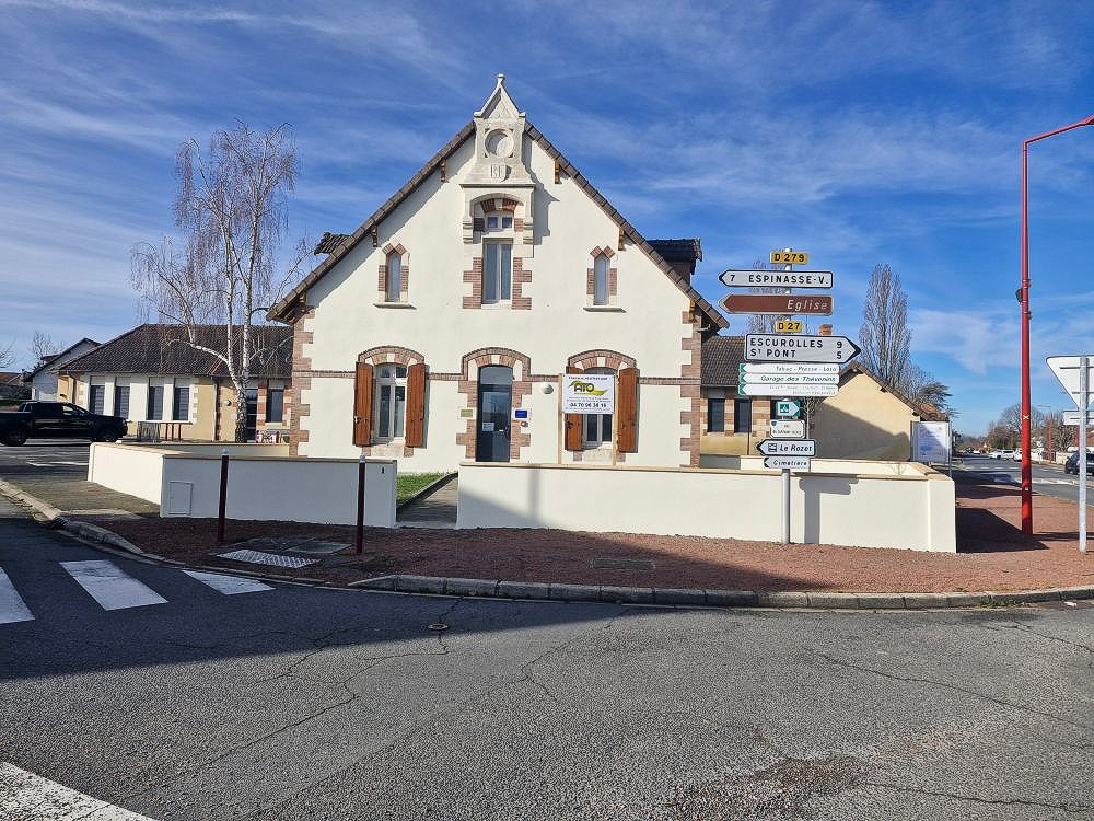 Un bâtiment blanc aux volets bruns et au toit à pignon se dresse derrière un muret, avec des panneaux de signalisation à proximité, par une journée ensoleillée.