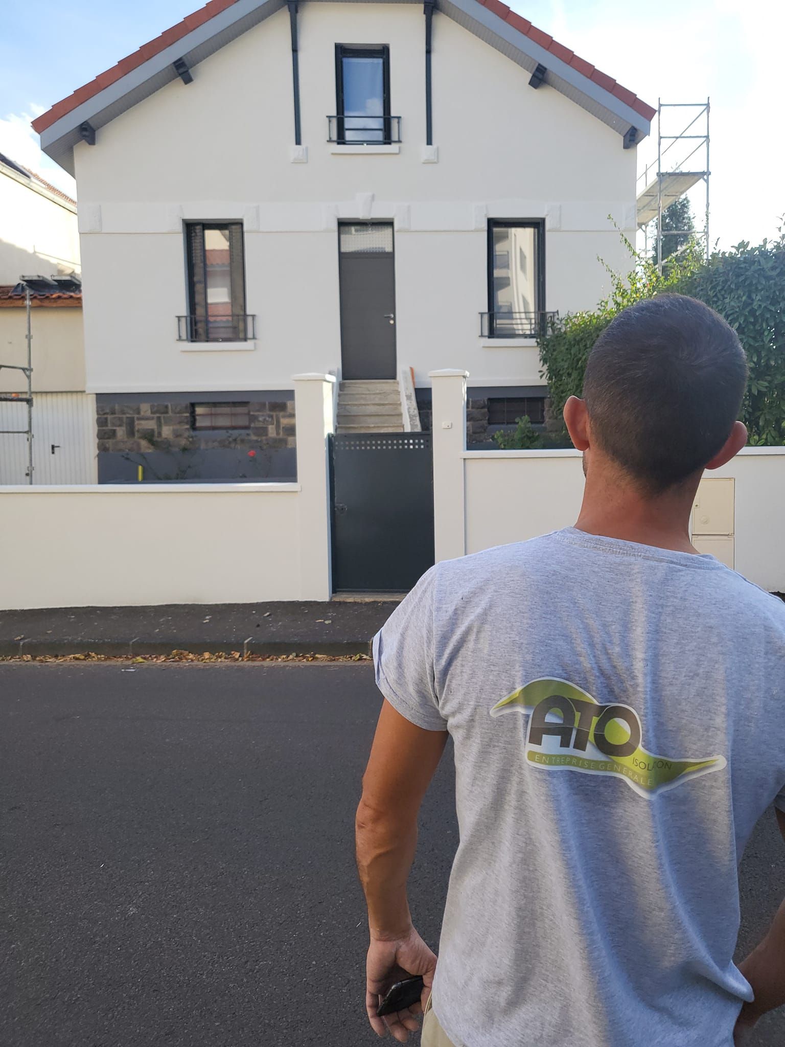 Une personne portant un t-shirt gris avec un logo se tient dans une rue face à une maison blanche à deux étages dont la porte d'entrée est surélevée.