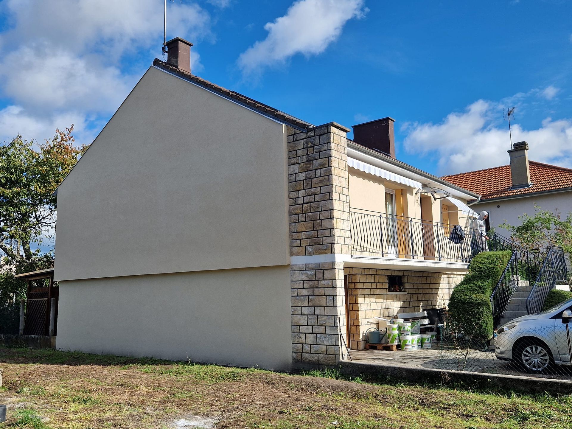 Une maison claire avec une façade en pierre, un balcon et une allée où une voiture est garée par une journée ensoleillée.