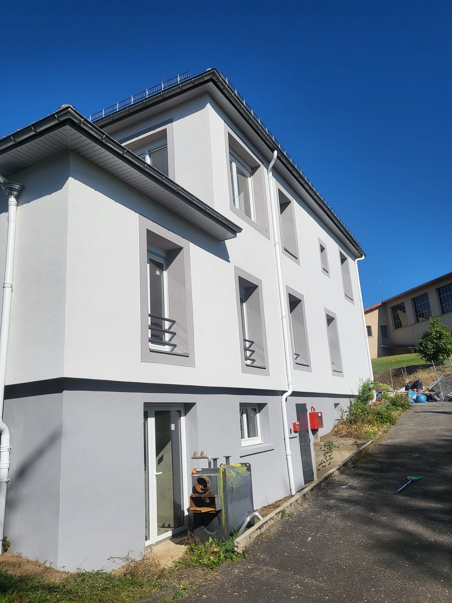 Un bâtiment de trois étages avec une façade gris clair, une base gris foncé et des descentes de gouttière blanches se détachant sur un ciel bleu clair.