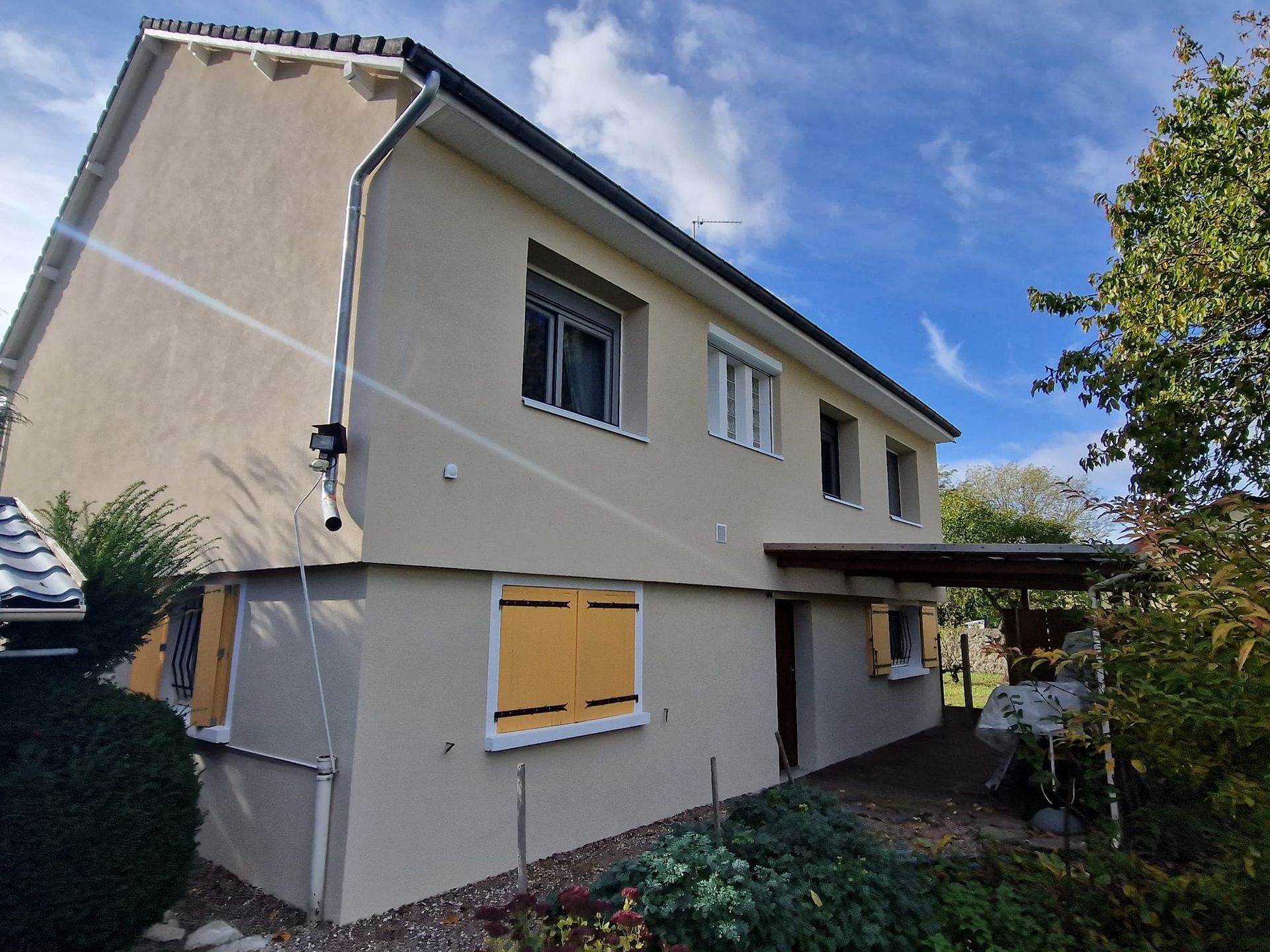 Une maison à deux étages aux murs en stuc beige, aux tuiles sombres et aux volets jaunes, sous un ciel bleu ensoleillé.