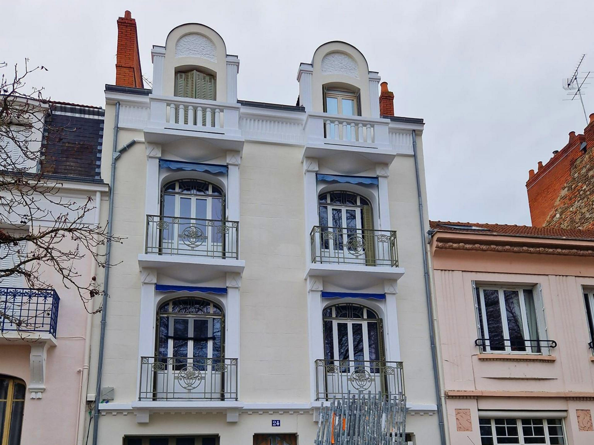 Un bâtiment historique de trois étages, de couleur jaune pâle, avec deux lucarnes symétriques et des balustrades de balcon décoratives en fer forgé.