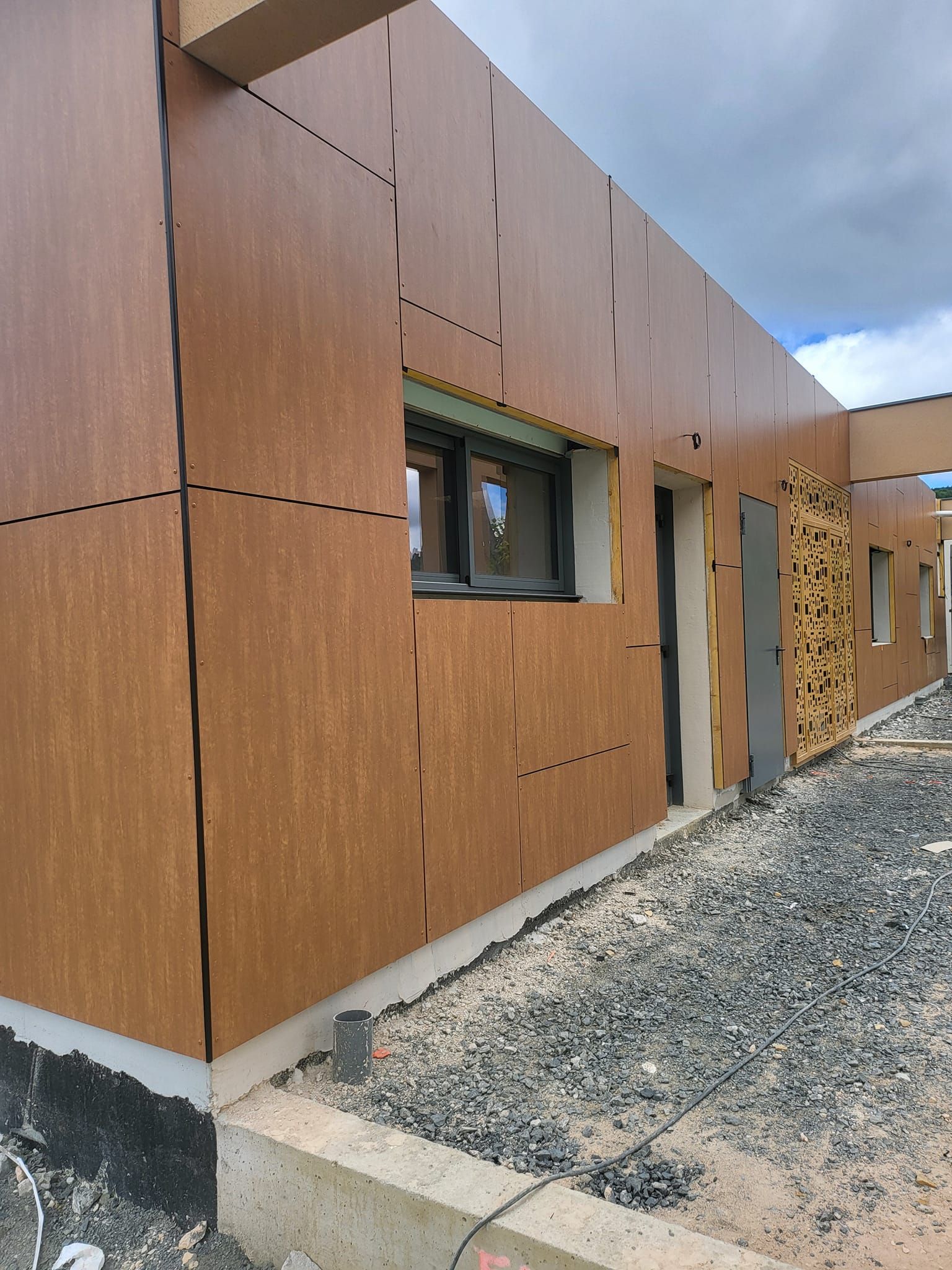 Façade d'un bâtiment en construction avec un bardage en panneaux bruns, une fenêtre, une porte et des panneaux décoratifs en treillis métallique.