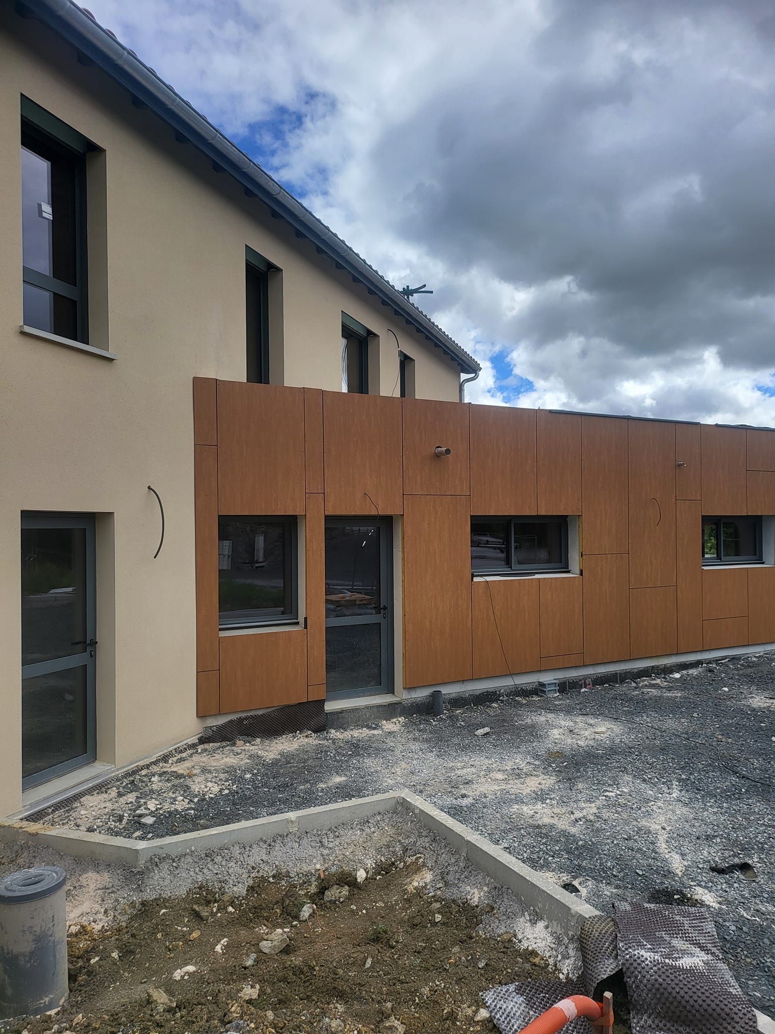Un bâtiment de deux étages en construction, avec un enduit beige et des lambris, des portes vitrées et une cour gravillonnée.
