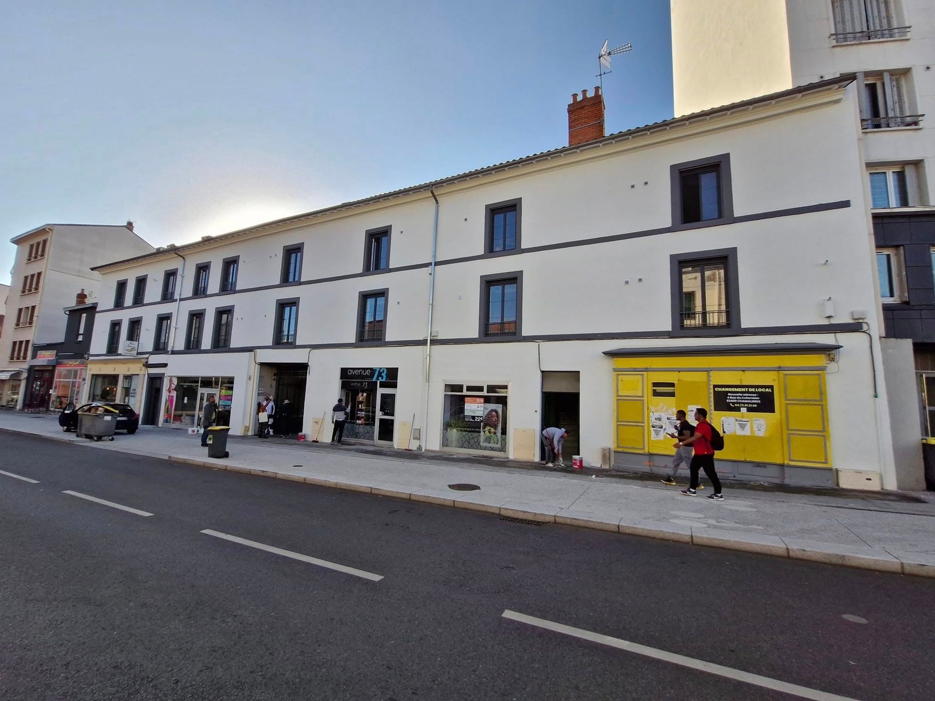 Un long bâtiment blanc de deux étages avec des devantures de magasins au rez-de-chaussée, dont une avec une façade jaune proéminente.